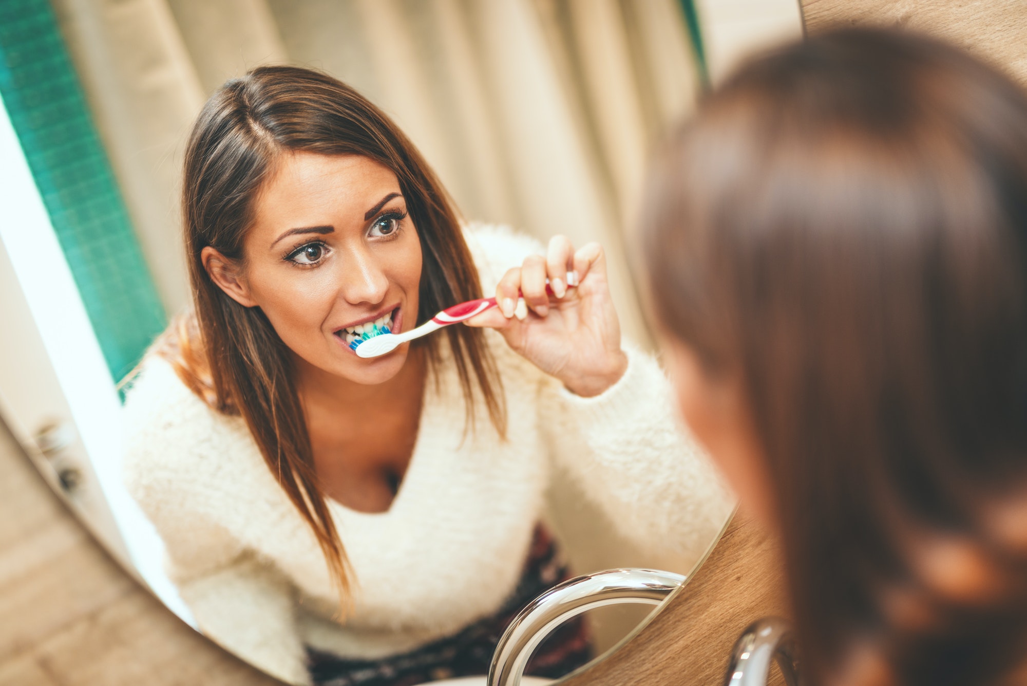 Smiling woman brushing teeth in mirror, emphasizing oral health's role in overall wellness - Des Moines Dental Associates.